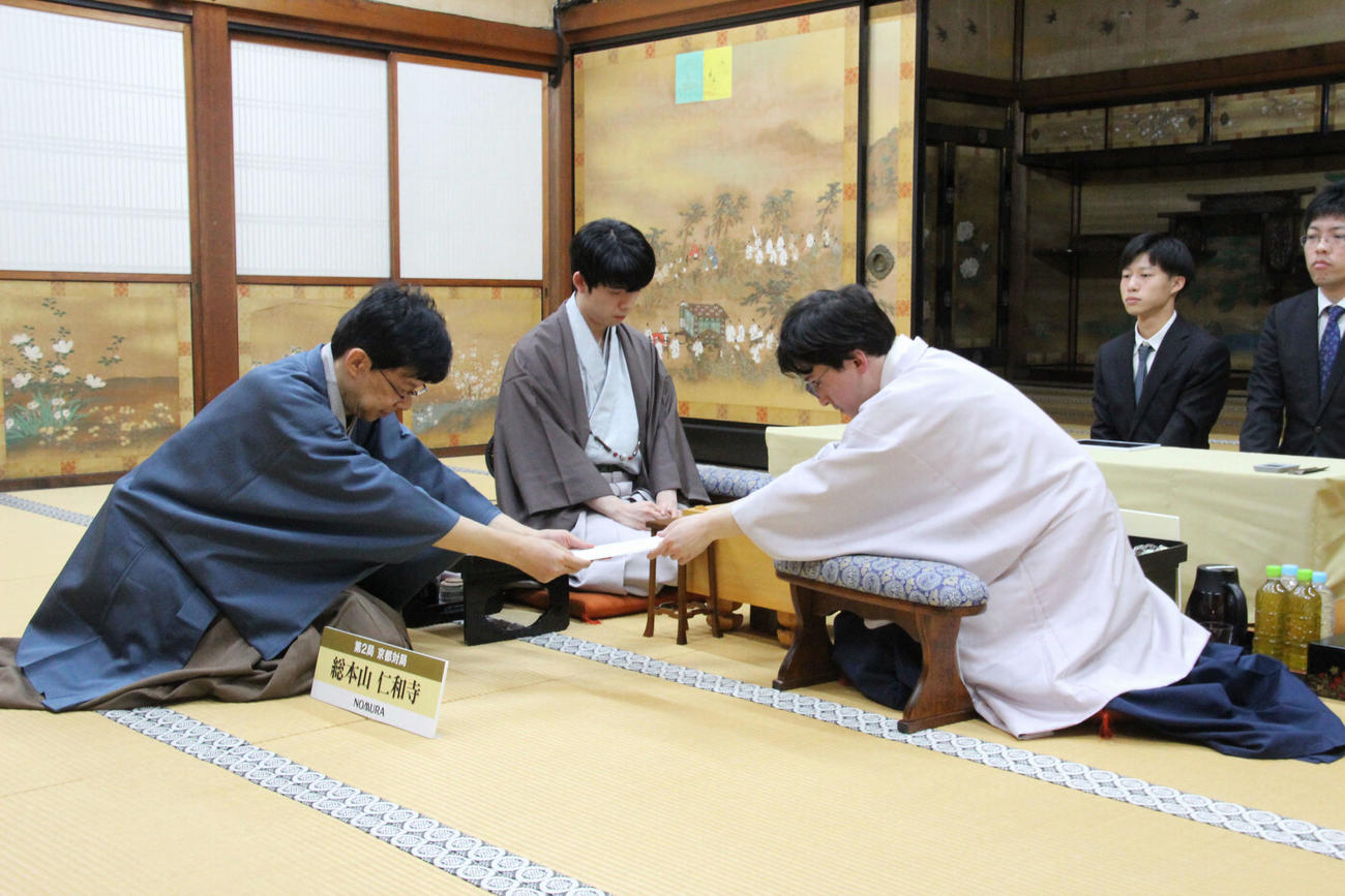立会人の谷川浩司17世名人（左）に封じ手を手渡す伊藤匠七段（右）、中央は藤井聡太8冠（（日本将棋連盟提供）
