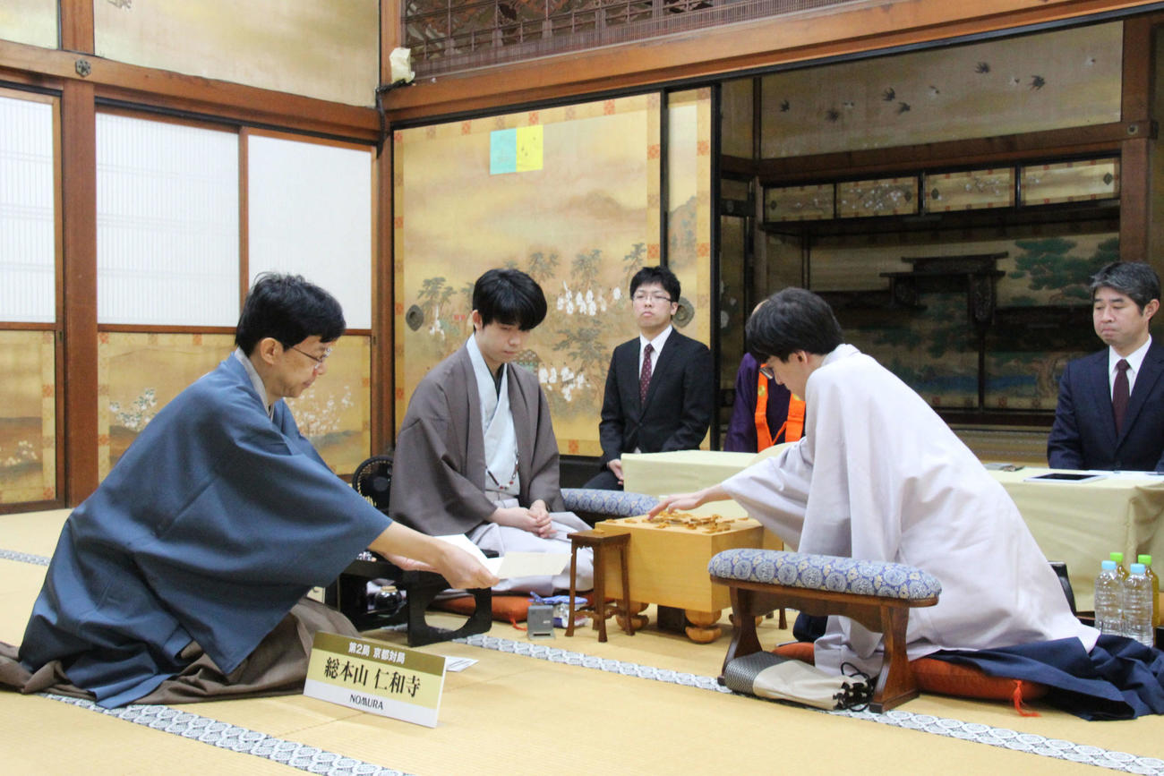 立会人の谷川浩司17世名人（左）が手に持つ封じ手を確認する藤井聡太8冠（左）、封じ手を指す伊藤匠七段（日本将棋連盟提供）