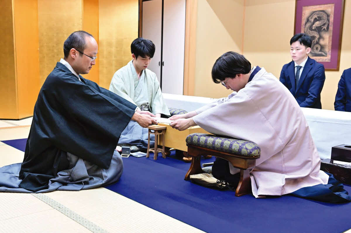 立会人の渡辺明九段（左）に封じ手の入った封筒を渡す挑戦者の伊藤匠七段（右）。中央は藤井聡太竜王（日本将棋連盟提供）