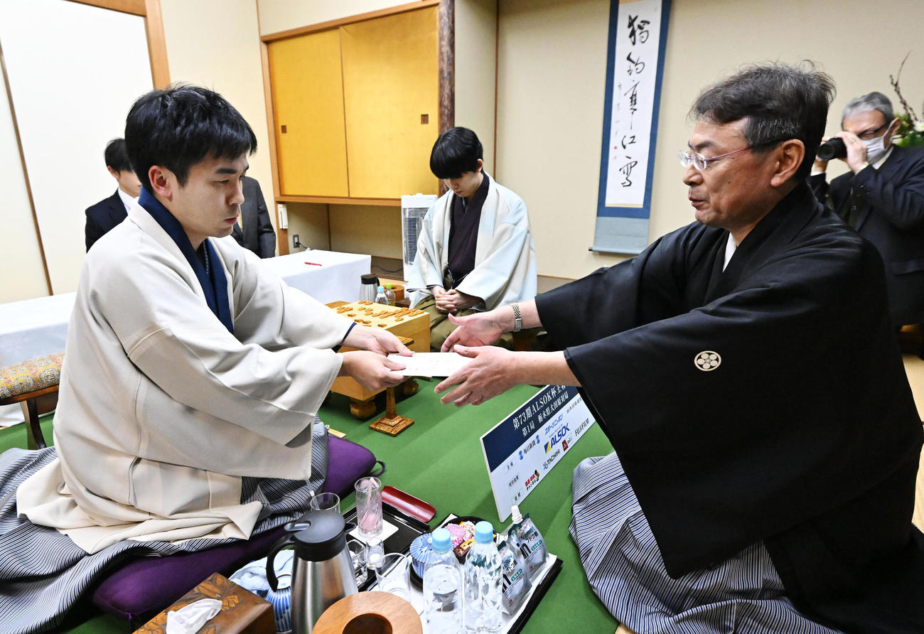 立会の塚田泰明九段（手前右）に封じ手を渡す挑戦者の菅井竜也八段。中央奥は藤井聡太王将（代表撮影）
