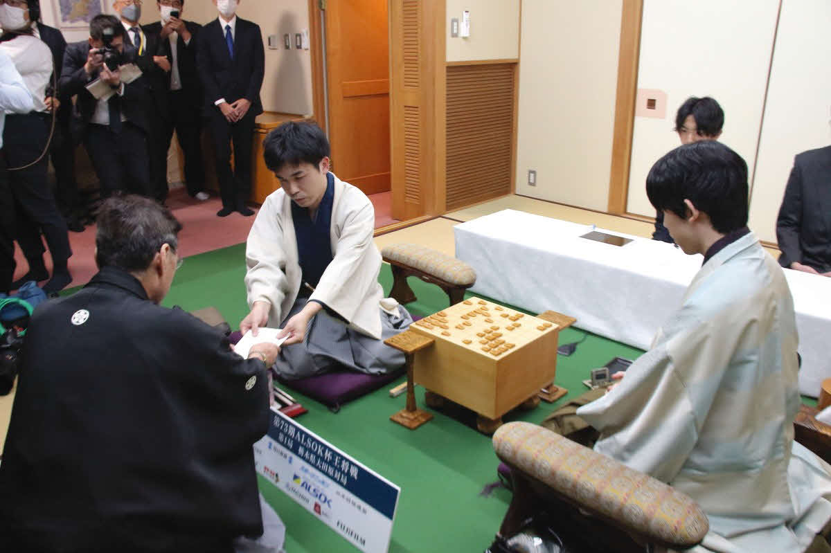 菅井竜也八段（左）は立会人の塚田泰明九段に封じ手の入った封筒を手渡す（右は藤井聡太王将、日本将棋連盟提供）