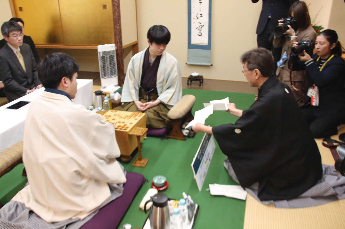 菅井竜也八段（左）の封じ手を開封した立会人の塚田泰明九段（右）が藤井聡太王将（中央）に見せて対局は再開（日本将棋連盟提供）