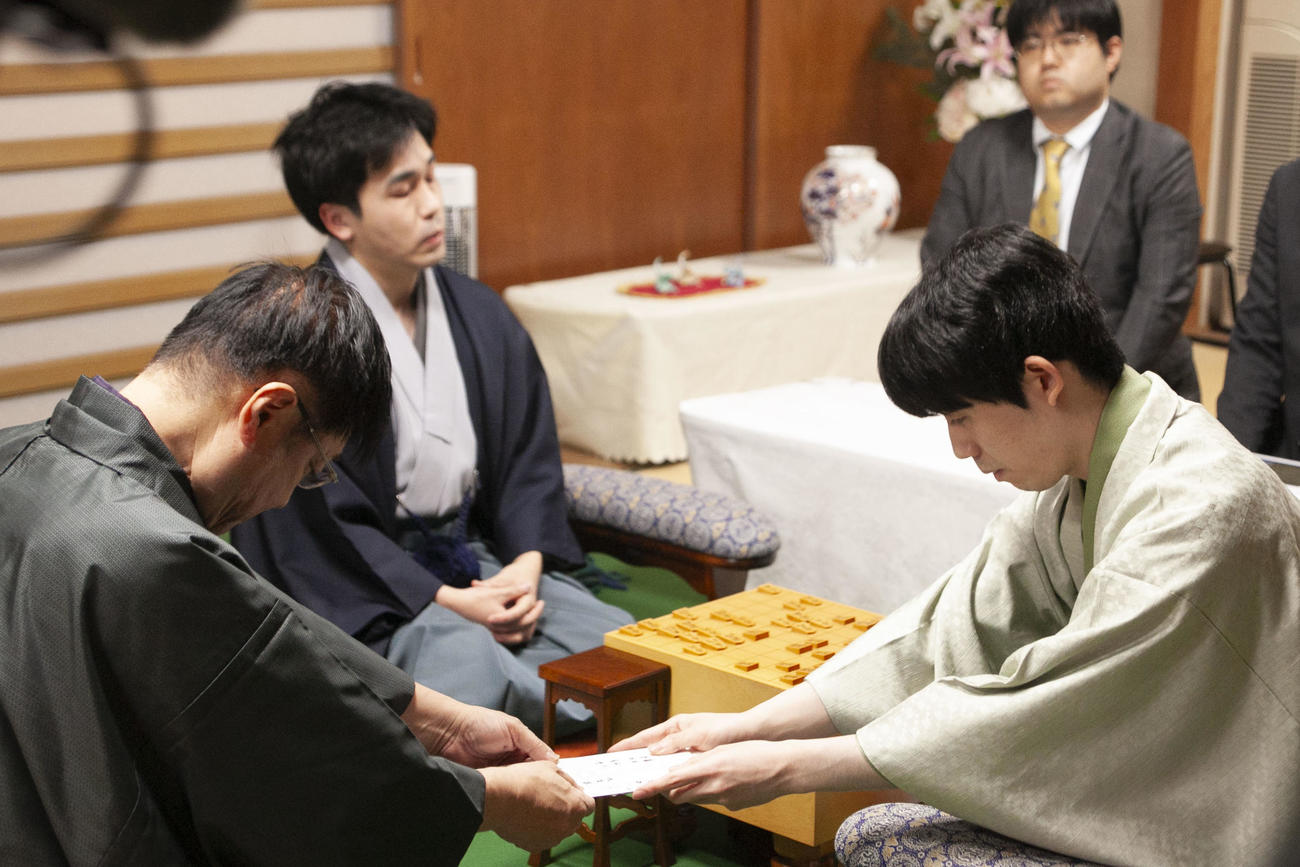 封じ手の封筒を立会人の小林健二九段に手渡す藤井聡太王将（右）、菅井竜也八段（左）（日本将棋連盟提供）