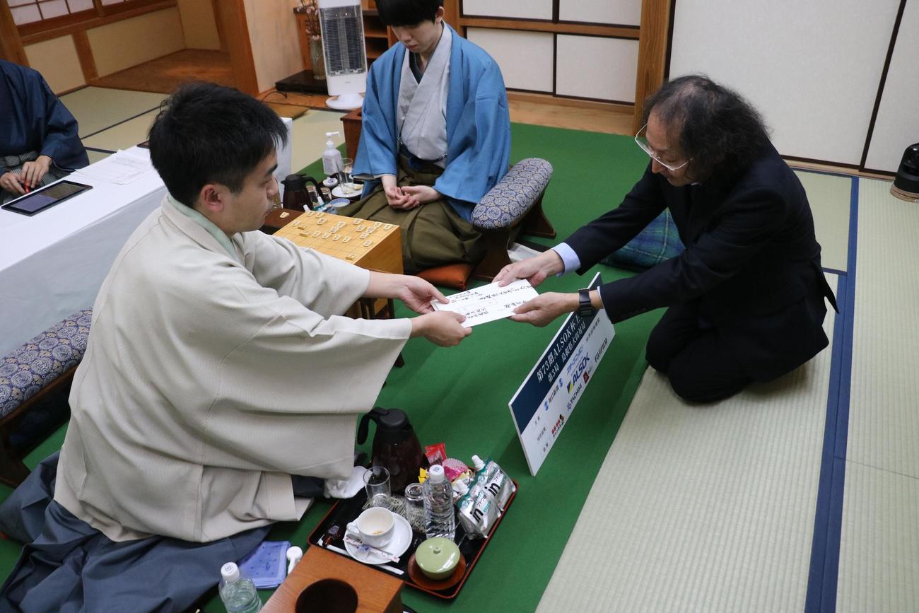 封じ手の封筒を立会人の福崎文吾九段（右）に手渡す菅井竜也八段、中央は藤井聡太王将（日本将棋連盟提供）