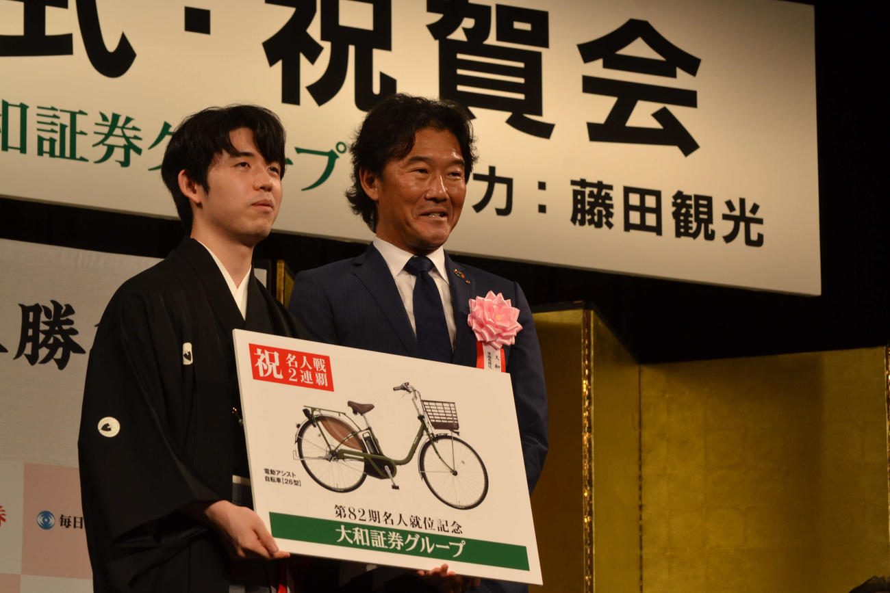 記念品に贈呈された電動アシスト自転車の目録を手にする藤井聡太名人（左）