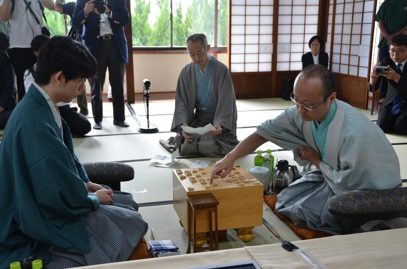 立会人の中田功八段（中央）が読み上げる封じ手を指す渡辺明九段（右）、藤井聡太王位（日本将棋連盟提供）