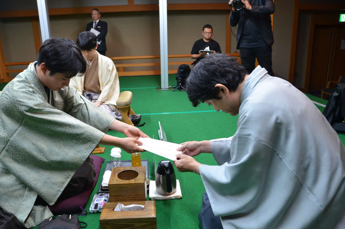 タイトル戦初登場となった佐々木勇気八段（左）は封じ手の入った封筒を立会人の森内俊之九段（右）に手渡す（奥は藤井聡太竜王、日本将棋連盟提供）