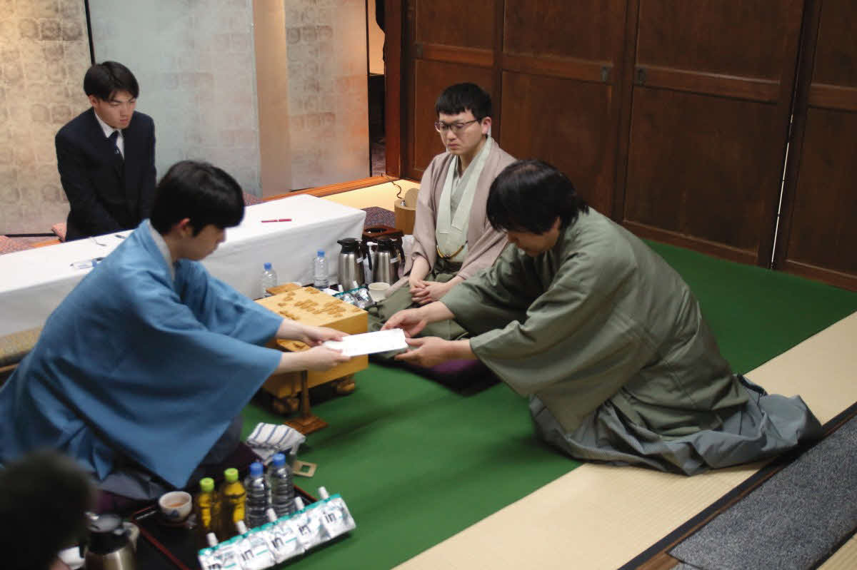藤井聡太王将（左）は立会人の藤井猛九段（右）に封じ手の入った封筒を渡す。中央は永瀬拓矢九段（日本将棋連盟提供）