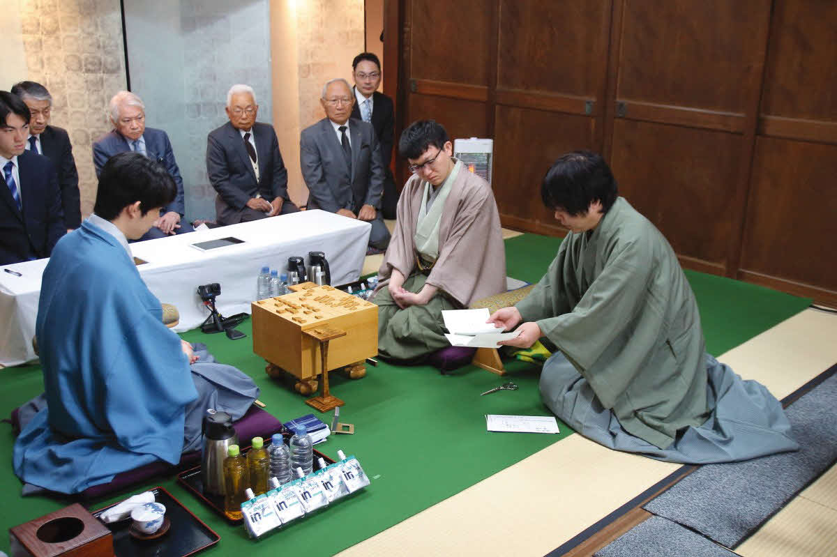 王将戦第5局で藤井聡太王将（左）の封じ手を開封する立会人の藤井猛九段（右）と確認する永瀬拓矢九段（右から2人目、日本将棋連盟提供）