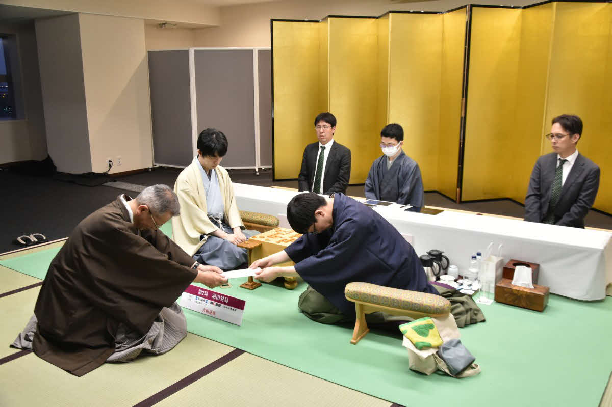 立会人の中村修九段（左）に封じ手の入った封筒を渡す永瀬拓矢九段（右、中央は藤井聡太名人日本将棋連盟提供）