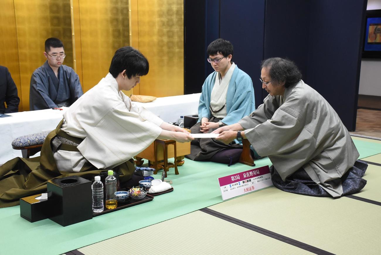 封じ手の封筒を立会人の福崎文吾九段（右）に手渡す藤井聡太名人（左）。中央は永瀬拓矢九段（日本将棋連盟提供）