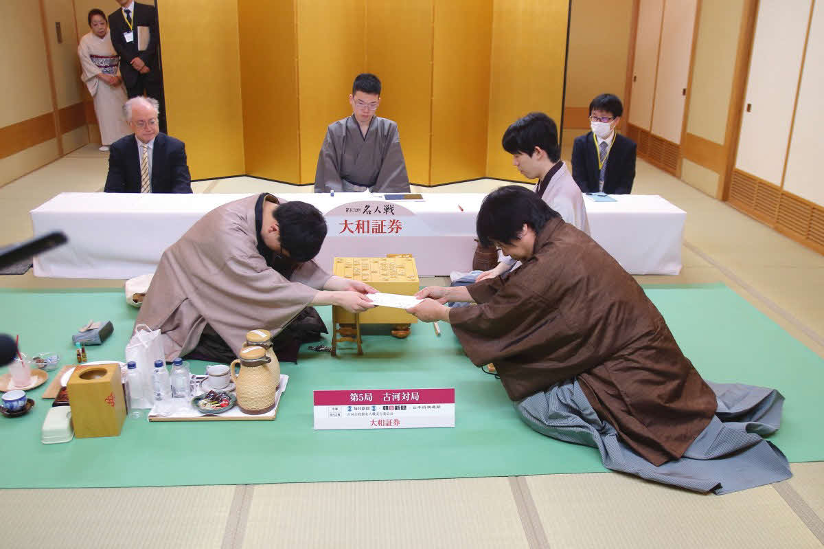 永瀬拓屋九段（左）は封じ手の入った封筒を立会人の藤井猛九段（右）に手渡す（奥は藤井聡太名人、日本将棋連盟提供）