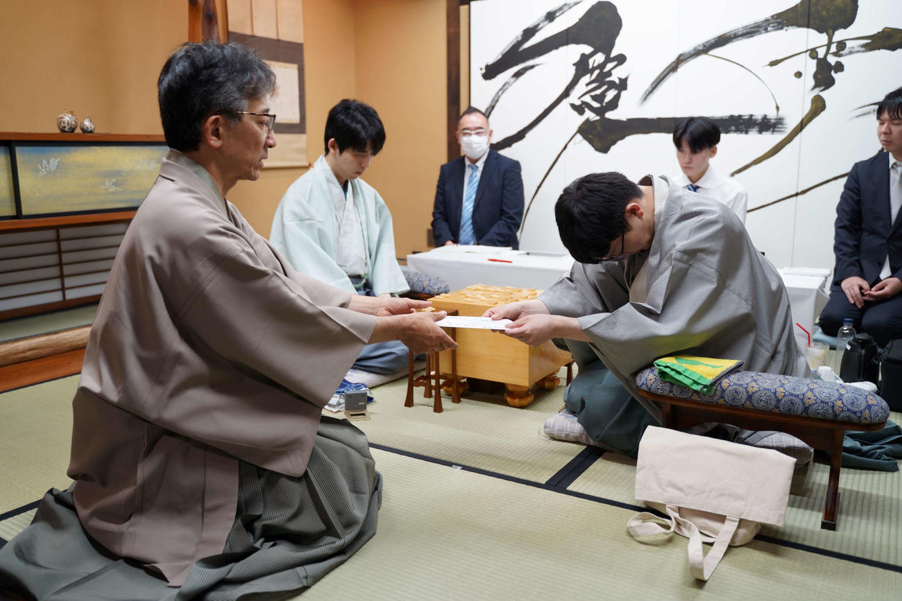 封じ手の封筒を立会人の佐藤康光九段（左）に手渡す永瀬拓矢九段（右）藤井聡太王位（中央）（日本将棋連盟提供）