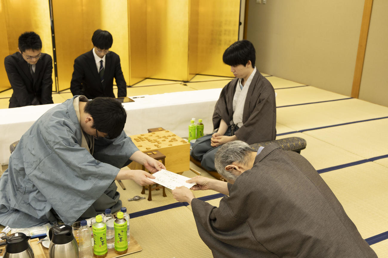 封じ手の封筒を立会人の中田功八段（中央）に手渡す永瀬拓矢九段（左）、藤井聡太王位（右）（日本将棋連盟提供）