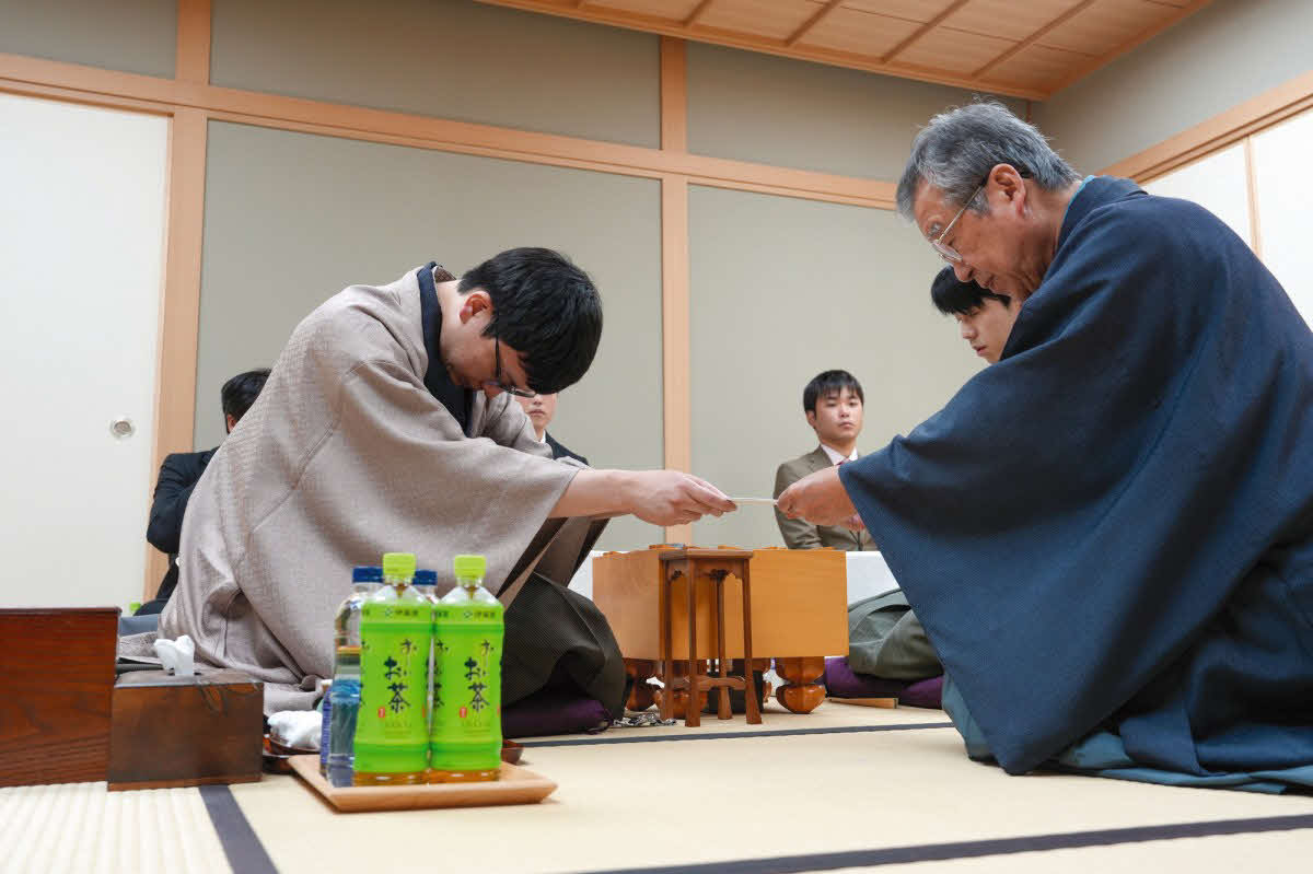 王位戦第6局は永瀬拓矢九段（左）が封じ手を立会人の青野照市九段（右）に渡して初日を終了（奥は藤井聡太王位、日本将棋連盟提供）