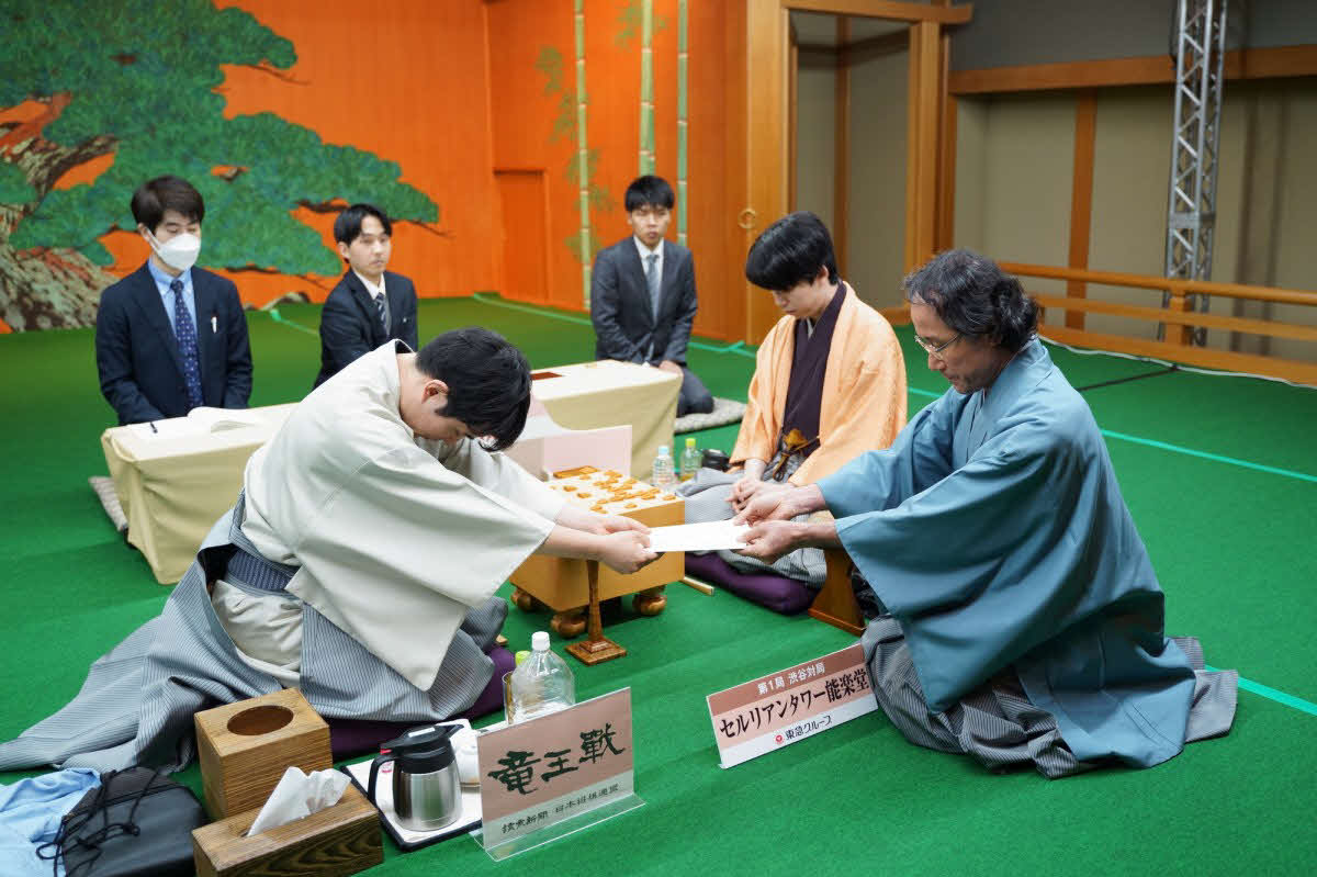 竜王戦第1局は佐々木勇気八段（左）が立会人の深浦康市九段（右）に封じ手の入った封筒を渡して初日を終える（中央は藤井聡太竜王、日本将棋連盟提供）