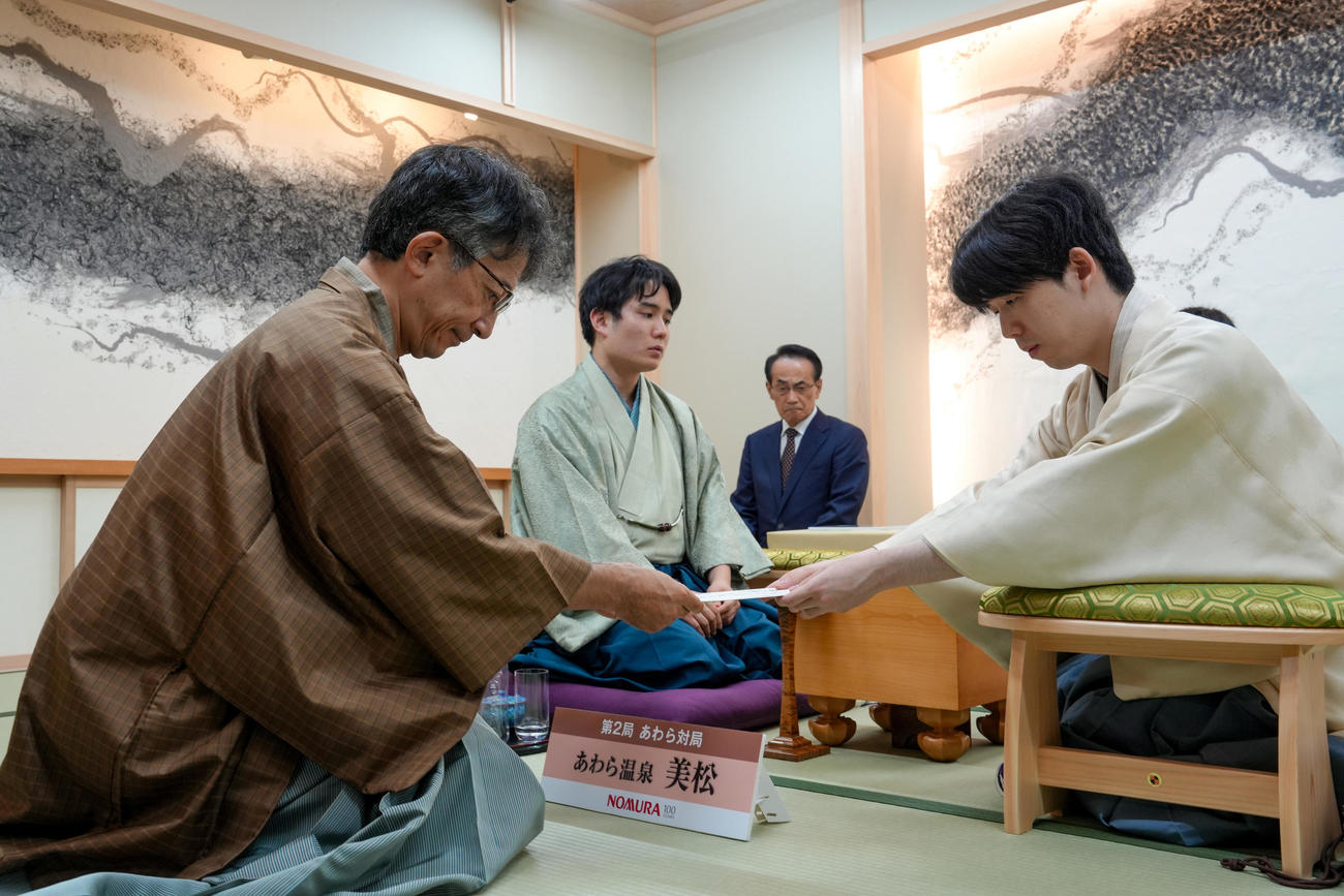 封じ手の封筒を立会人の佐藤康光九段（左）に手渡す藤井聡太竜王（右）、中央は佐々木勇気八段（日本将棋連盟提供）