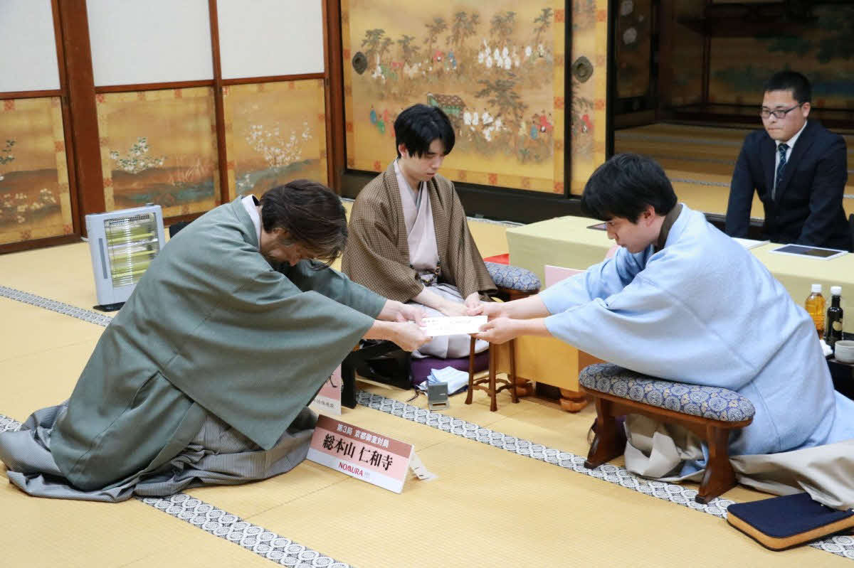 立会人の久保利明九段（左）に佐々木勇気八段（右）が封じ手の入った封筒を渡して竜王戦7番勝負第3局の初日は終了（中央は藤井聡太竜王、日本将棋連盟提供）