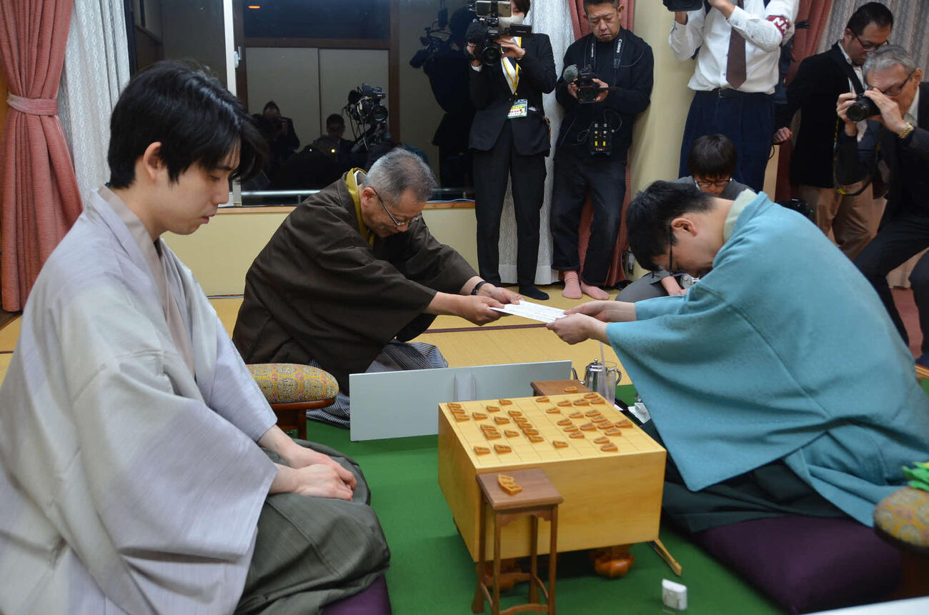 王将戦第5局初日は永瀬拓矢九段（右）が封じ手を立会人の中村修九段（中央）に渡して終了（左は藤井聡太王将、日本将棋連盟提供）