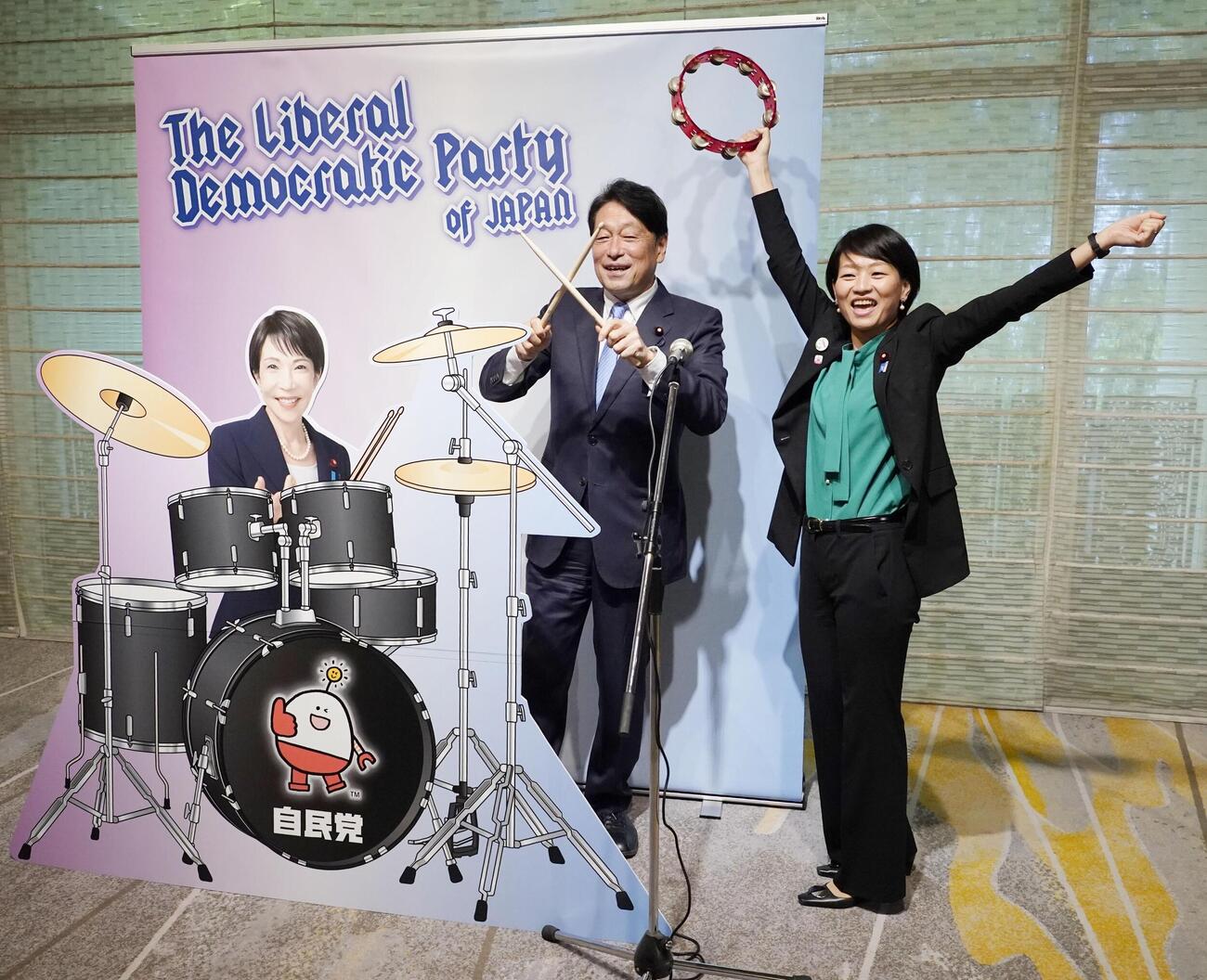 自民党大会の会場に設置された高市首相のフォトパネルの横でポーズをとる小野寺五典税調会長（左）と鈴木貴子広報本部長（共同）
