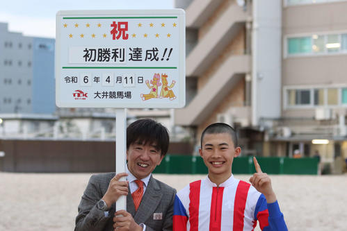 初勝利を挙げ「イチバーン」ポーズを取る川崎の佐野遥久騎手（右）と「どっきり」のようにプラカードを持つ大井の須田和伸調教師