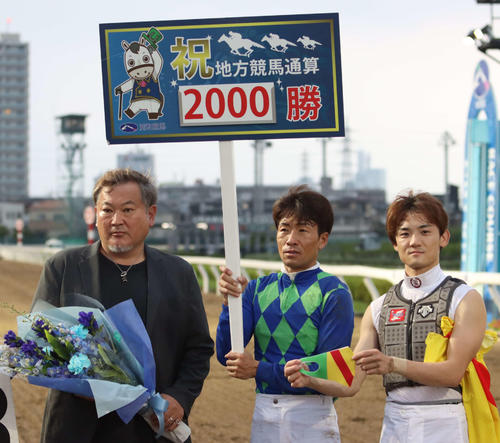 地方競馬通算2000勝を達成した浦和の小久保智調教師（左）と左海誠二元騎手の騎手服柄マスクを持つ笹川翼騎手（右）、プラカードを持つ森泰斗騎手