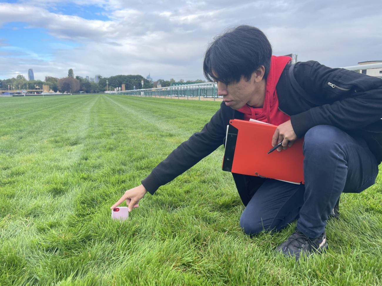 28日、潜入したパリロンシャン競馬場で芝の深さを見る奥田記者。スマホは「iPhone　SE」