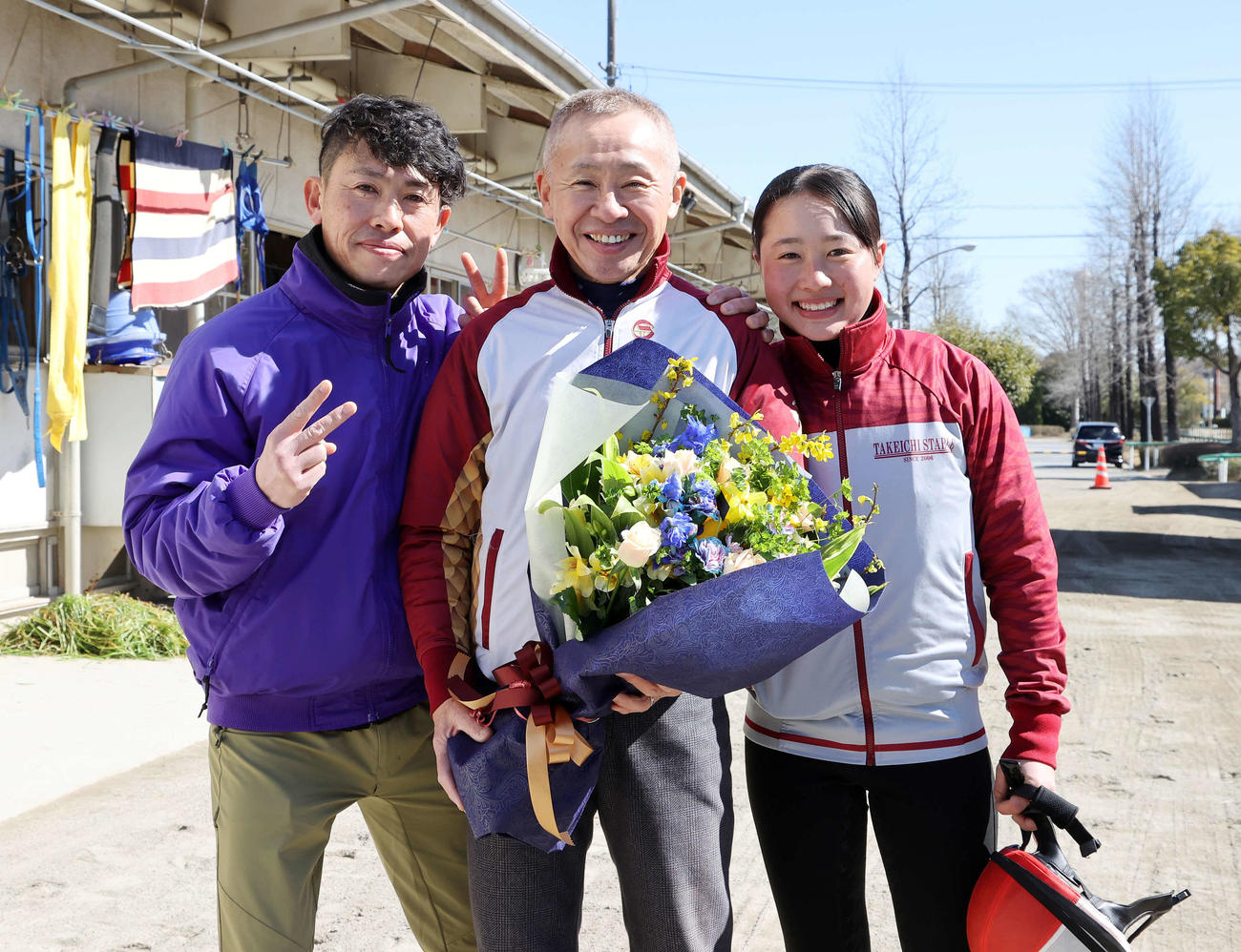 競馬学校騎手課程に在籍する孫の比呂さん（右）から花束を受け取り笑顔を見せる大江原調教師。左は長男の勝氏（撮影・鈴木正人）