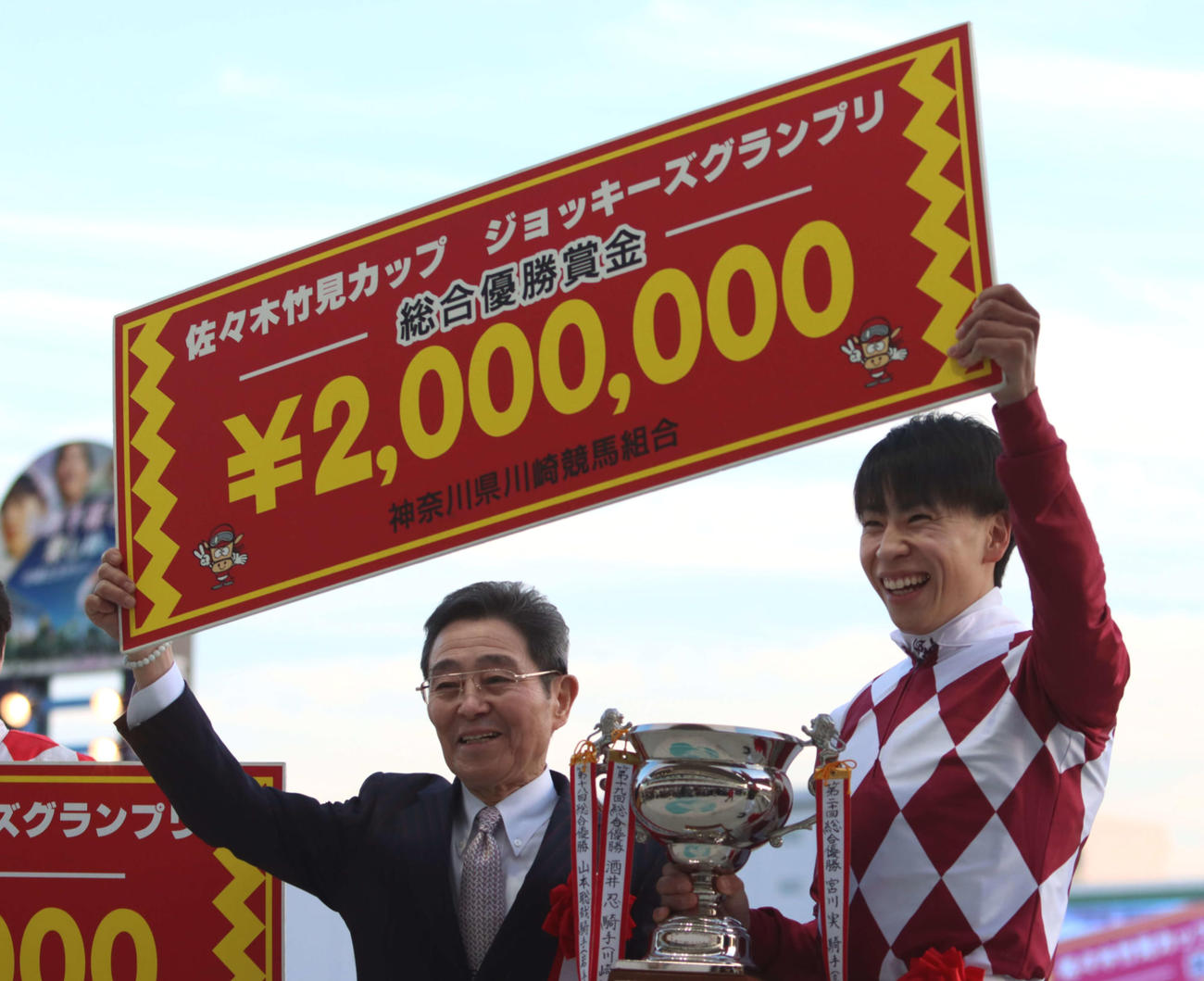 優勝し笑顔で賞金ボードを掲げる横山武史騎手（右）とプレゼンターの佐々木竹見元騎手
