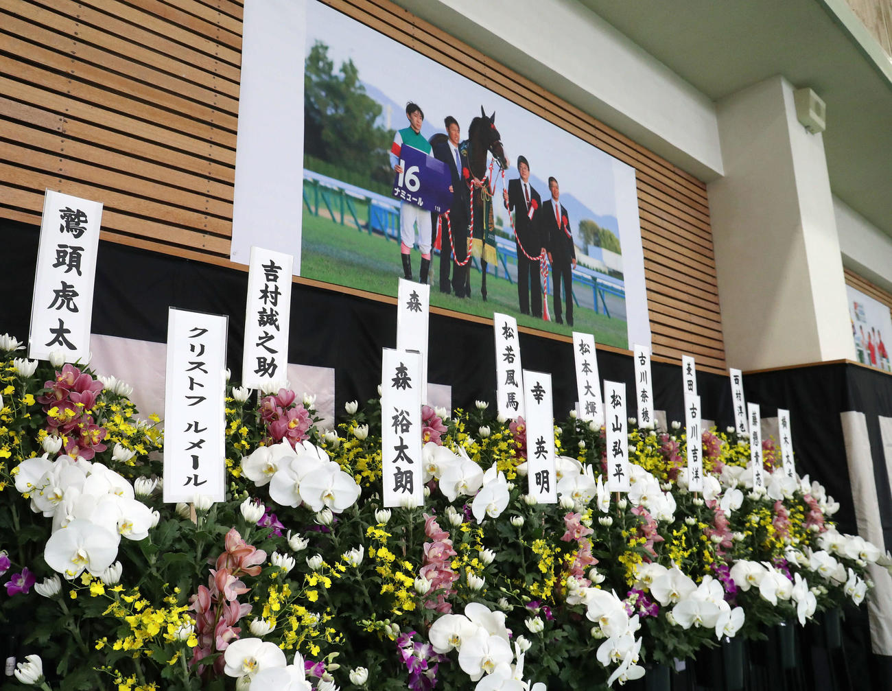 故藤岡康騎手の合同葬で騎手仲間から贈られた花。壁には昨年のマイルCSをナミュールで制した際の写真が飾られていた（撮影・白石智彦）