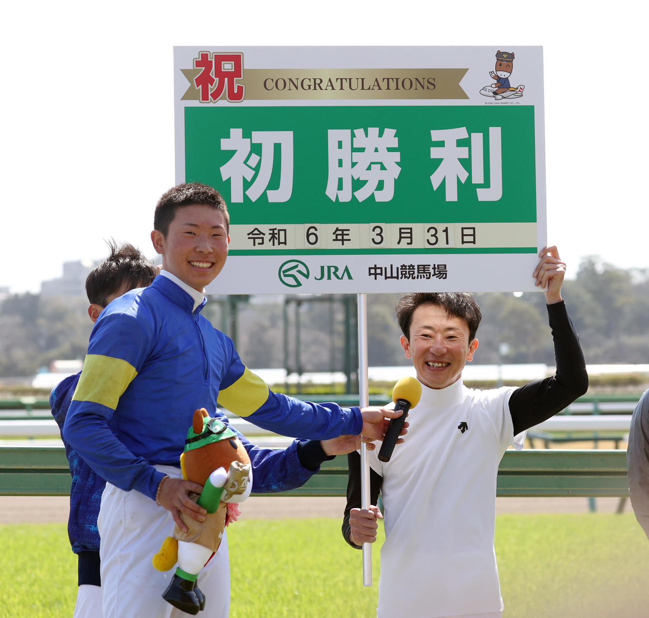 2024年3月、JRA初勝利を挙げた石神深道騎手（左）はボードを持つ父石神深一騎手にマイクを向ける