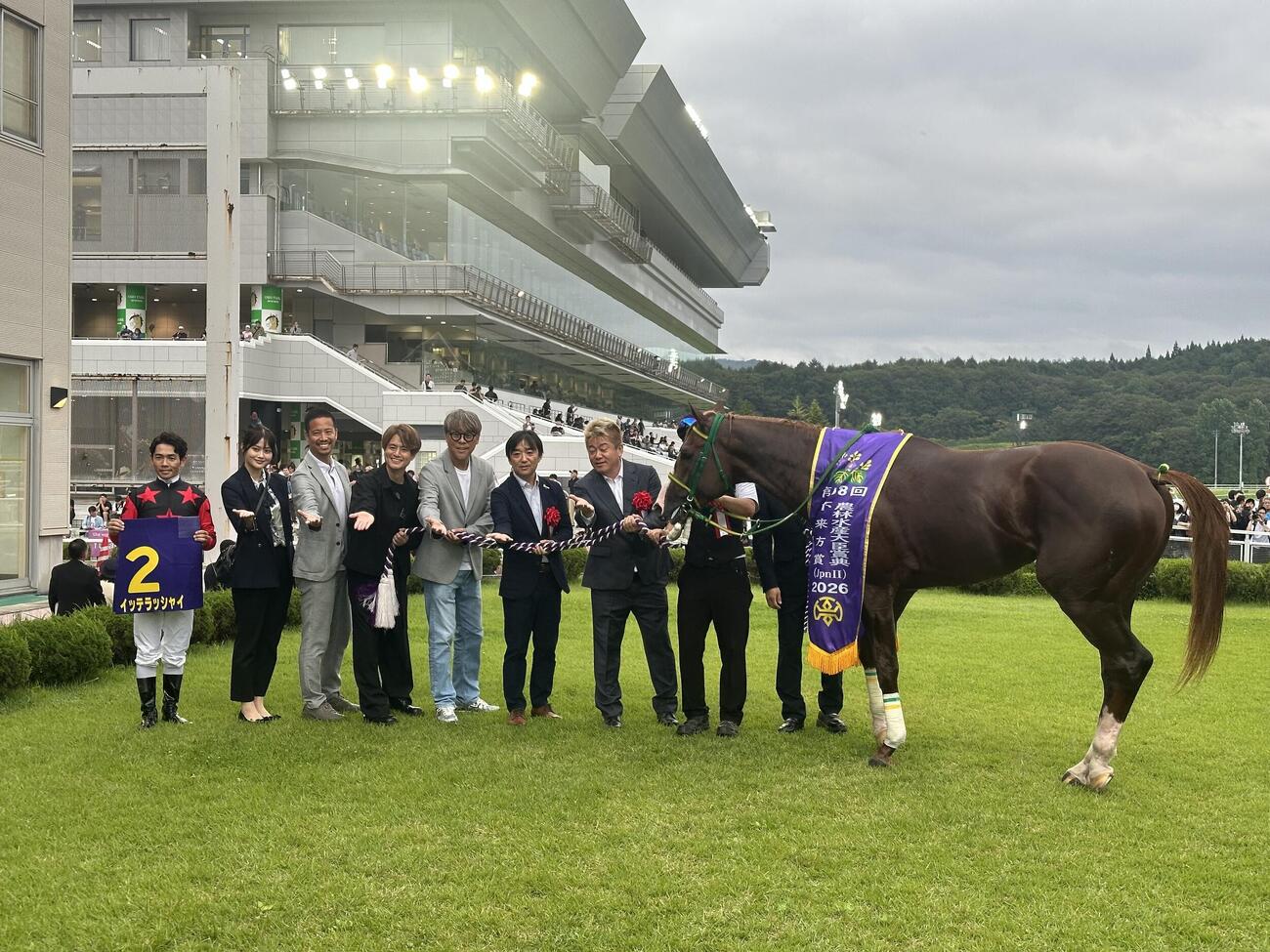 愛馬イッテラッシャイの横で「行ってらっしゃい」ポーズをする堀江貴文氏ら関係者（隣が斎藤誠師、左端が戸崎騎手）（撮影・松田直樹）