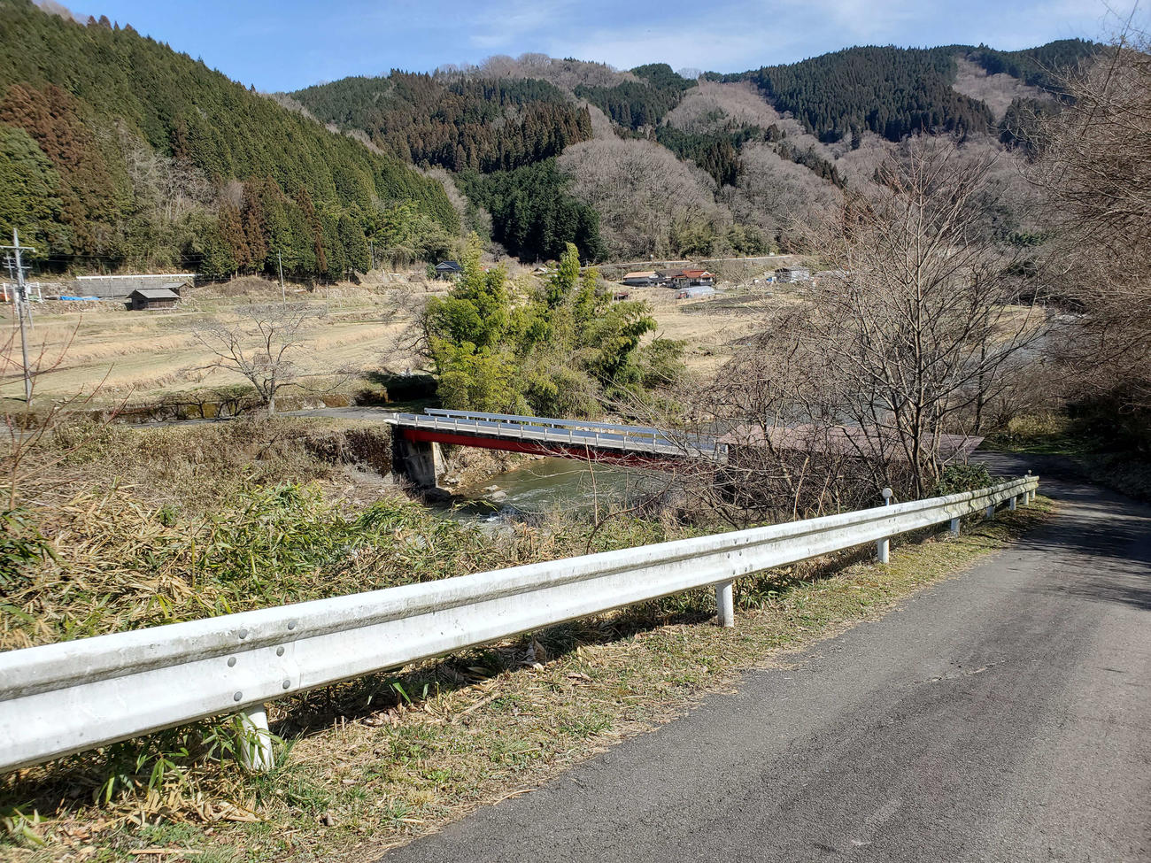 〈5〉駅から坂を下りて川を渡るといくつかの民家がある。県道は山沿いのかなたを通る