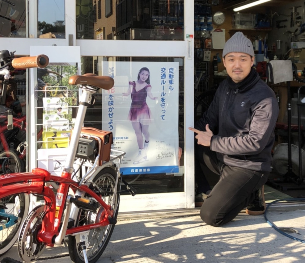 神戸市内の自転車屋「BIG　MOUNTAIN」の店頭で坂本花織のポスターと共に写真に納まる村山千春さん（村山さん提供）