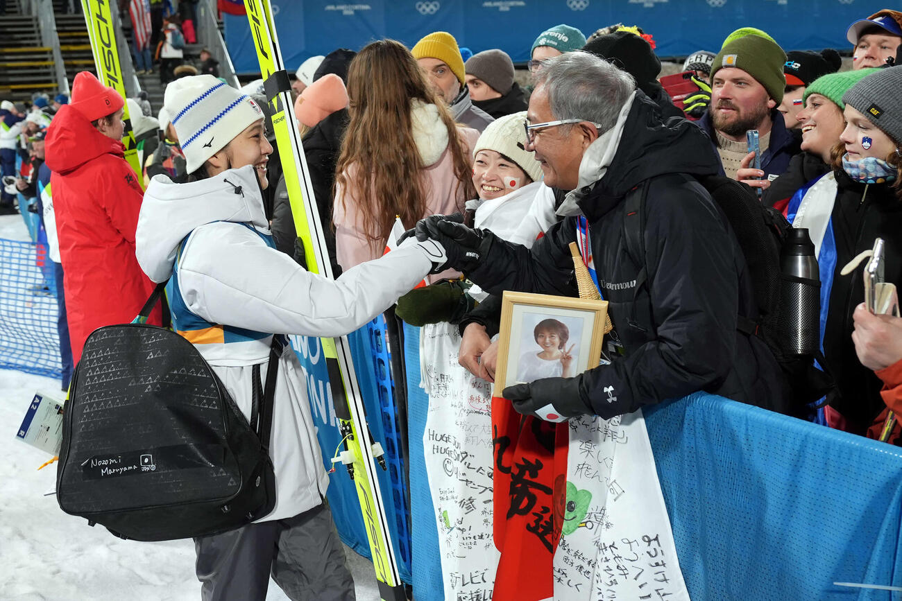 女子個人ラージヒルを終え、母信子さんの写真を持つ父守さん（右）と笑顔で握手する丸山希（撮影・パオロ　ヌッチ）