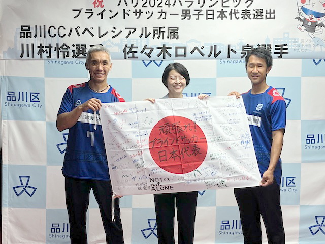 寄せ書き入りの日の丸を手にするパリパラリンピックのブラインドサッカー男子日本代表選手たち。左から佐々木、森沢恭子区長、川村