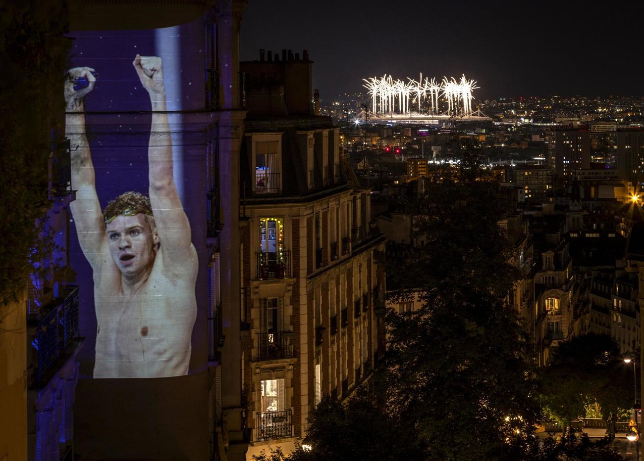 パリ五輪の閉会式が行われたフランス競技場に打ち上がる花火（右上）。左は映し出された競泳フランス代表のレオン・マルシャン（ゲッティ＝共同）