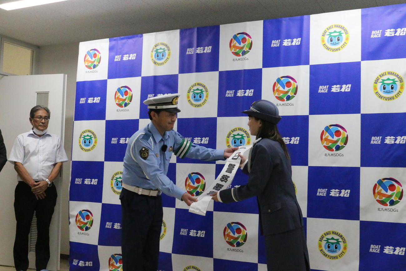 25日から31日までの飲酒運転撲滅週間を控え、若松警察署の一日署長に任命された松尾怜実（右）が、井町由起夫・若松警察署長からたすきを受け取る