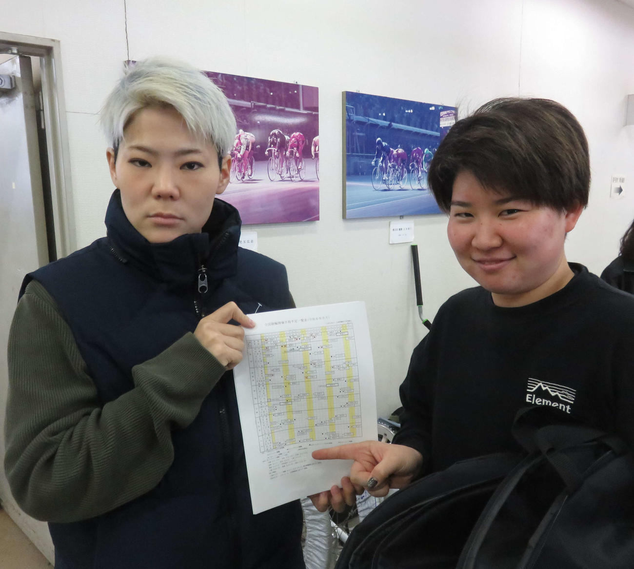 立川競輪　橋本佳耶（左）が九州久留米に冬季移動する畠山ひすいとともに熊本競輪の再開が記された開催日程に見入る