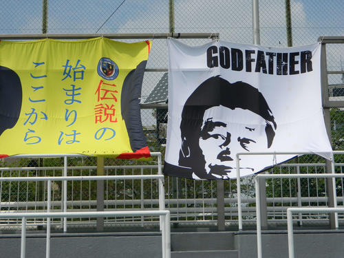 スタンドで風になびく東京ユナイテッドFCの旗。スポンサー企業会長の顔がイラストで描かれた
