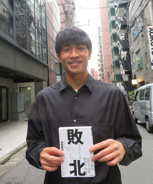 自著「敗北のスポーツ学」を手に新宿の街角に立つ井筒さん