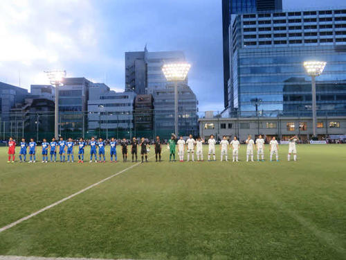 都心のビル群に囲まれたピッチで、試合前に整列する東京ユナイテッド（左側）と南葛SCのメンバー