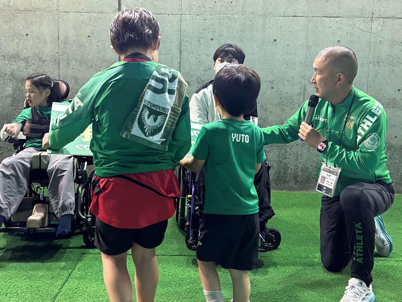 サッカー教室に参加した子供からグリーン・ハート・ルームの招待パネルを手渡される観戦者。右端が中村一昭さん