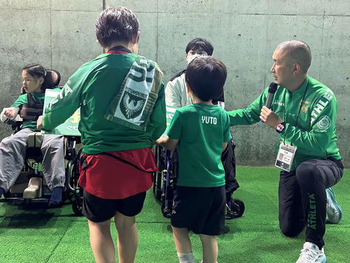 サッカー教室に参加した子供からグリーン・ハート・ルームの招待パネルを手渡される観戦者。右端が中村一昭さん