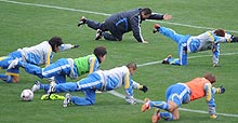選手とともに補強運動をする岡田監督（中央）。他の選手とは形が違うような…