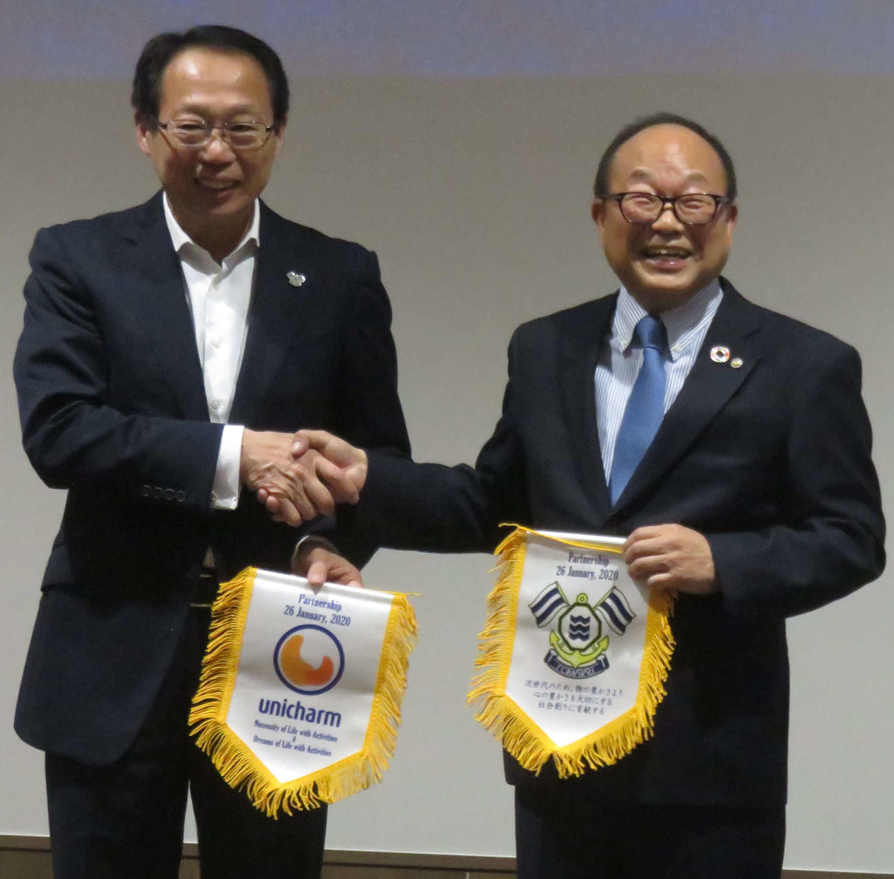 FC今治のシーズン方針発表会で今季のトップパートナーとなったユニ・チャームの石川副社長(右)とフラッグを交換する岡田オーナー