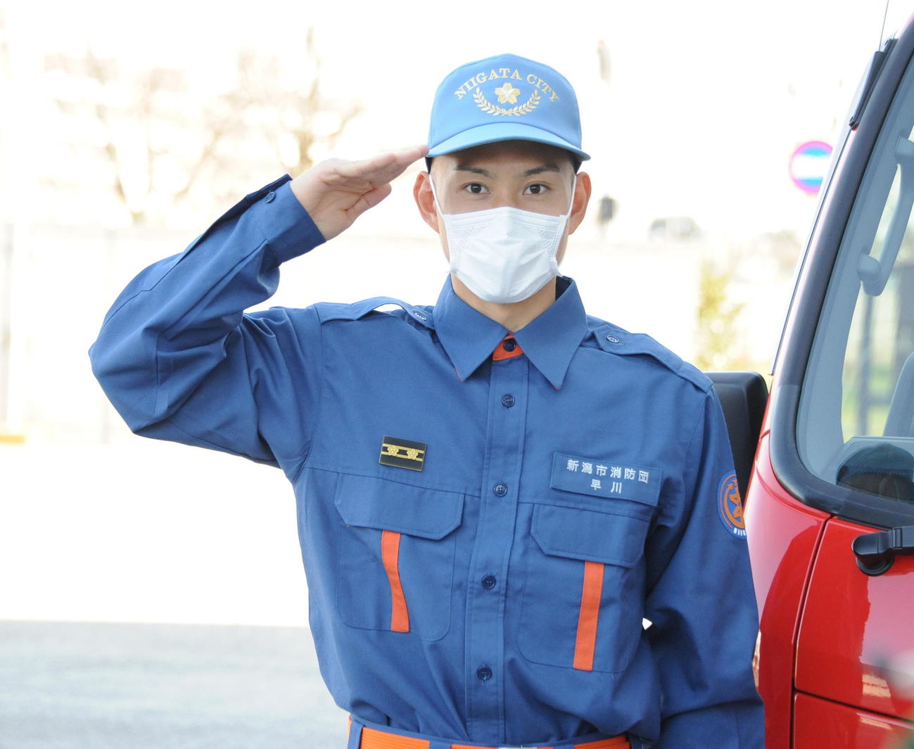 「1日消防団員」に任命された早川が消防車の前で敬礼ポーズ