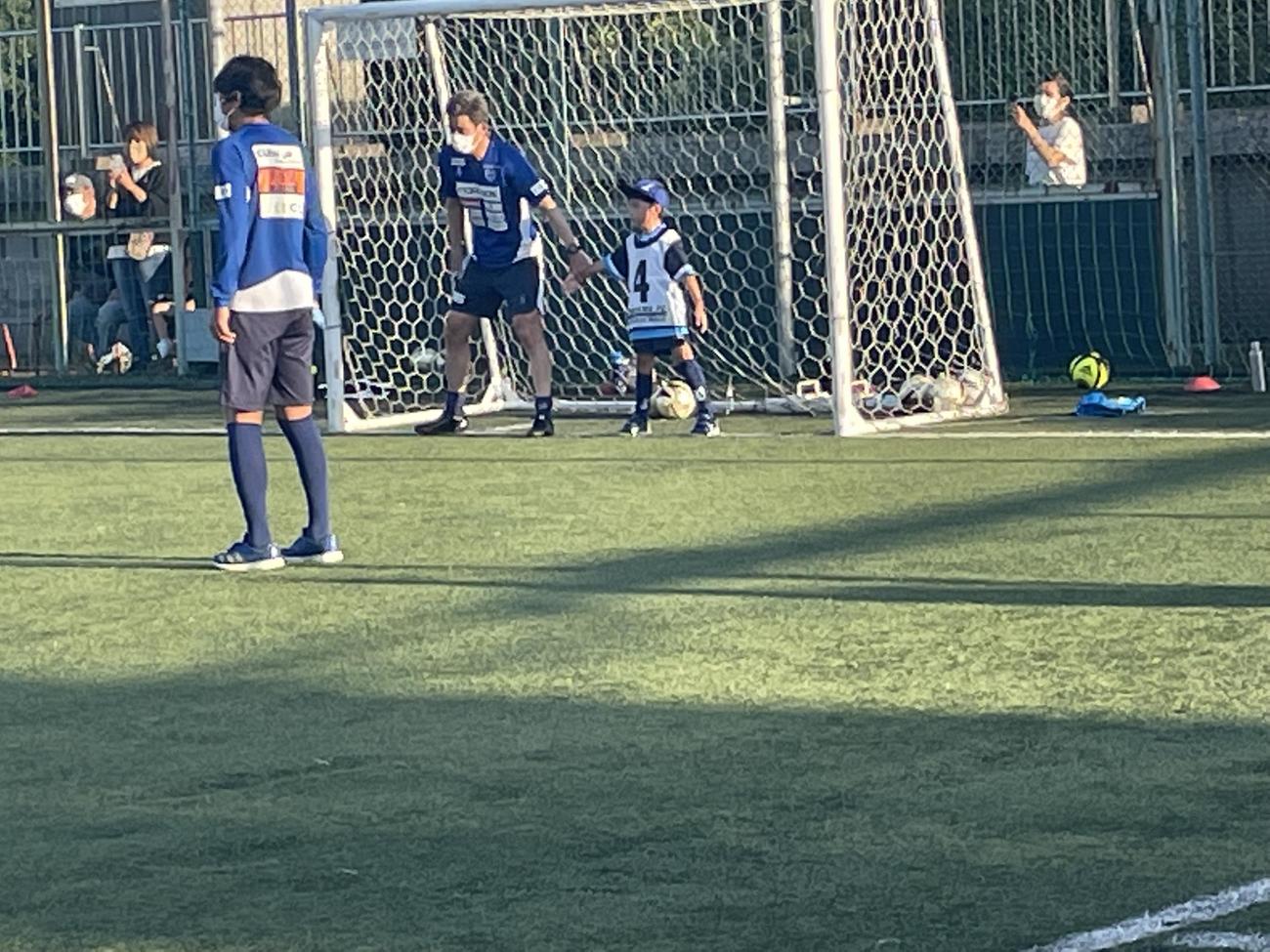 子どもと手をつなぎ、GKとしてゴールを守る横浜FCのFWカズ（三浦知良）と、ボールを待つMF中村（左）