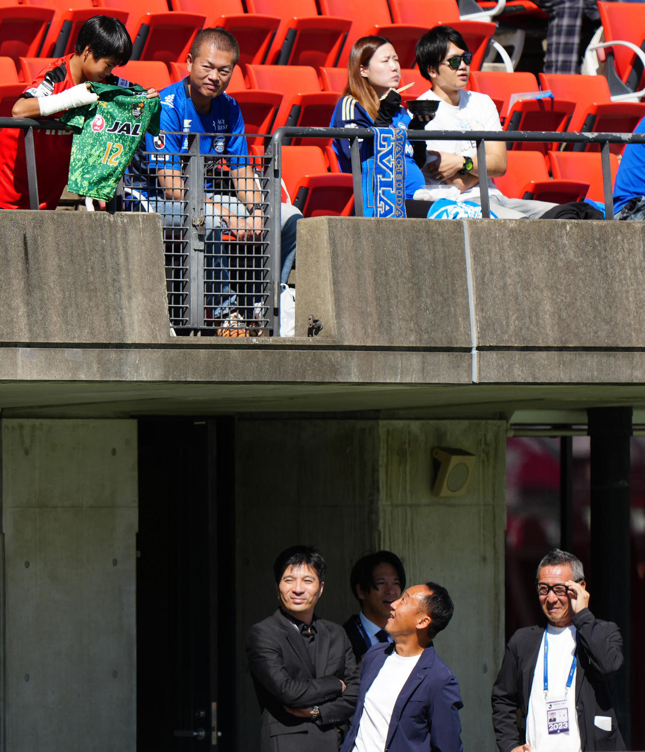 熊本対町田　試合前、スタンドで青森山田のユニホームを掲げる少年を笑顔で見つめる町田黒田監督（中央）。左は藤田社長（撮影・江口和貴）