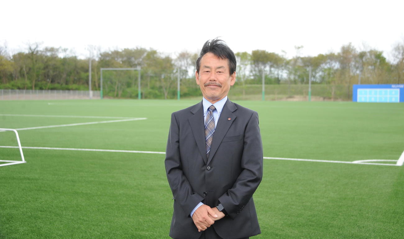新潟医療福祉大のキャンパス内に完成したサッカー場をバックに撮影に応じる同男子サッカー部佐熊監督